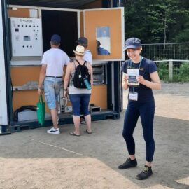 Tervetuloa tutustumaan Viljakkaaseen Okrassa 5.–8.7.2023! Viljakas-konttikuivurin edessä ihmisiä katsomassa sisään kuivuriin. Viljakas-esittelijä hymyilee ja katsoo kameraan esite kädessään.