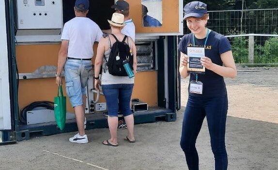 Viljakas-konttikuivurin edessä ihmisiä katsomassa sisään kuivuriin. Viljakas-esittelijä hymyilee ja katsoo kameraan esite kädessään.